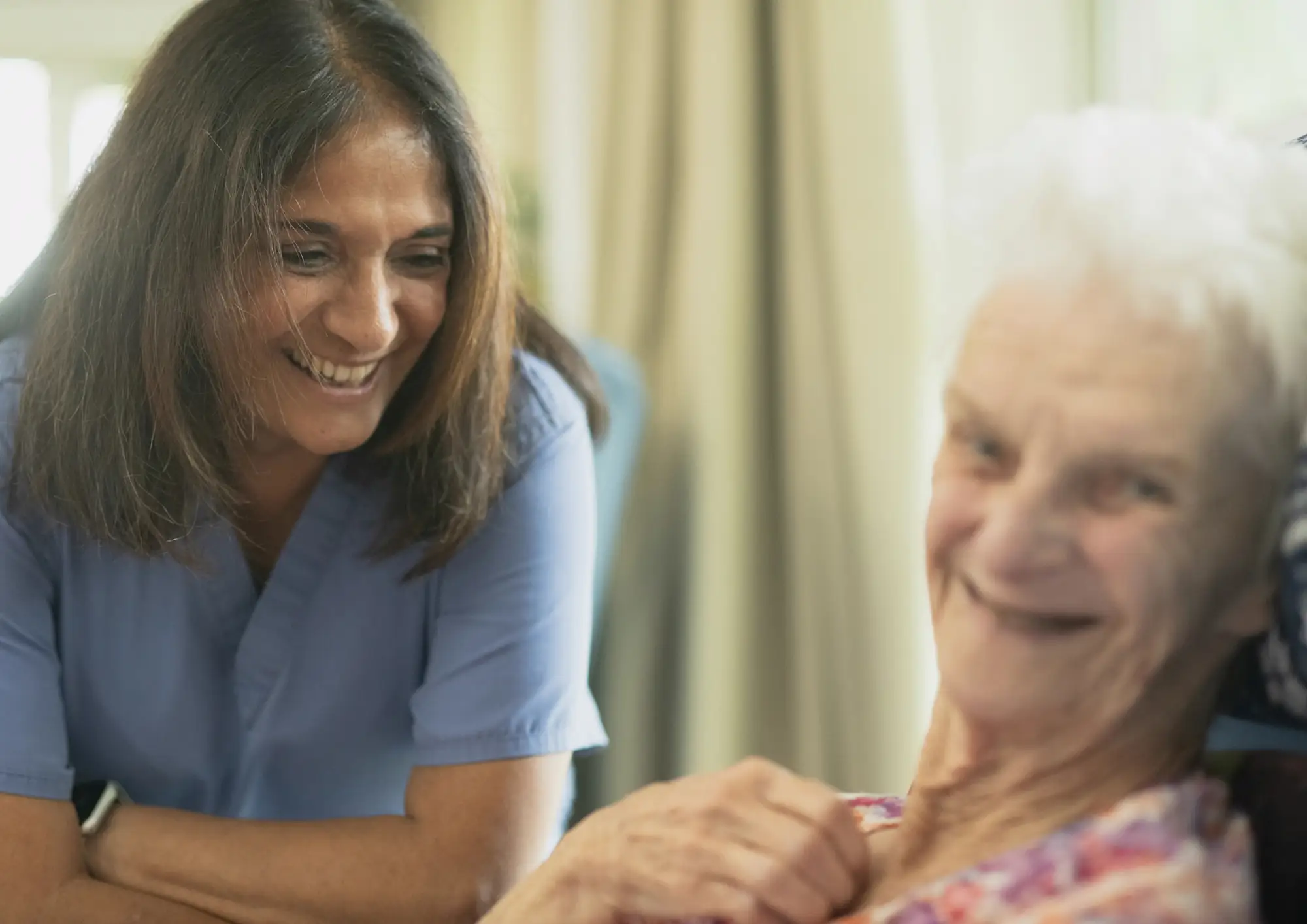 A smiling carer in blue scrubs talking with an elderly woman